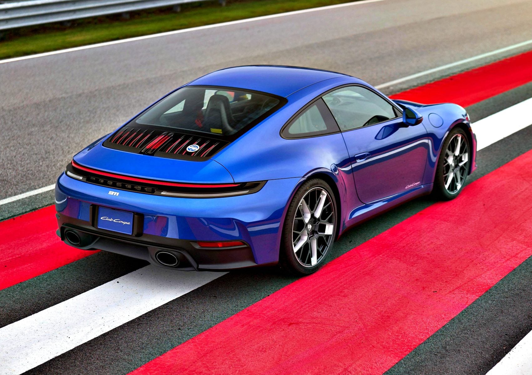 A blue Limited-Run 911 Carrera T Club Coupe sports car is parked on a red and white striped section of a racetrack, viewed from the rear left side.