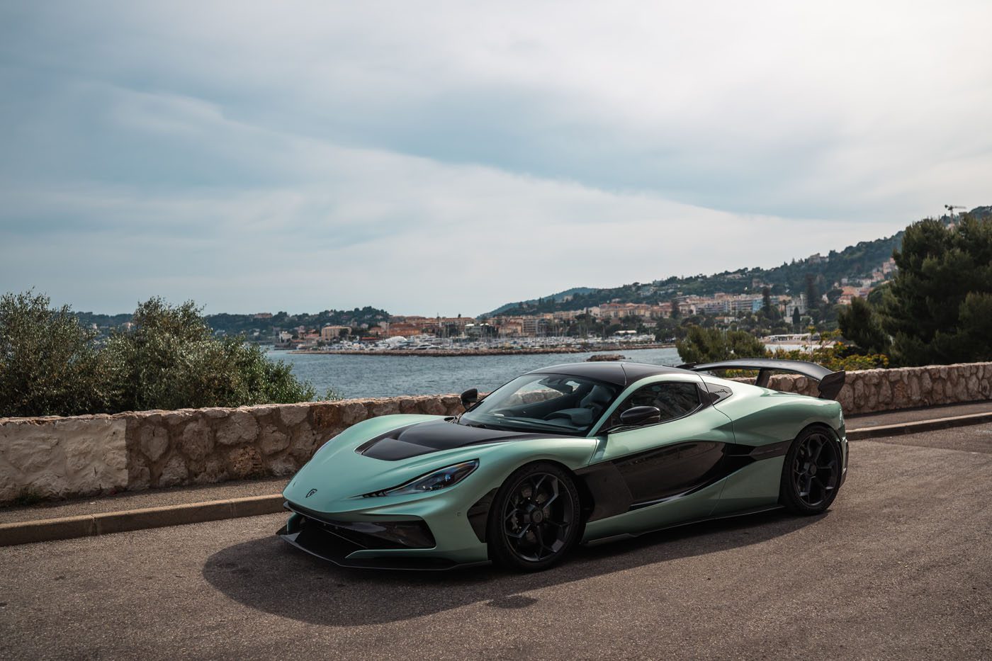 A green and black Rimac Nevera sports car is parked on a coastal road with a stone wall, overlooking the sea and a town with hills in the background, evoking the glamour of the Monaco Grand Prix.