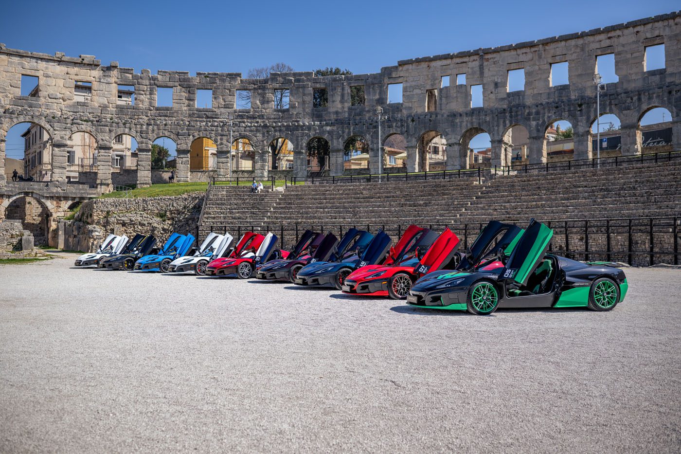 A lineup of Rimac Neveras with open scissor doors graces the front of an ancient coliseum, part of the exhilarating Trailblazers Tour, all under a clear blue sky.