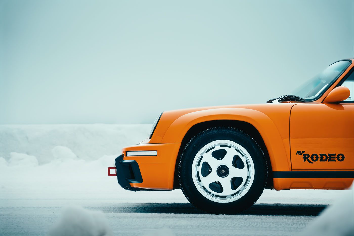 Front view of an orange sports car parked on a snowy landscape, featuring the black "RUF Rodeo" logo on its side—a unique display reminiscent of I.C.E. St. Moritz elegance.
