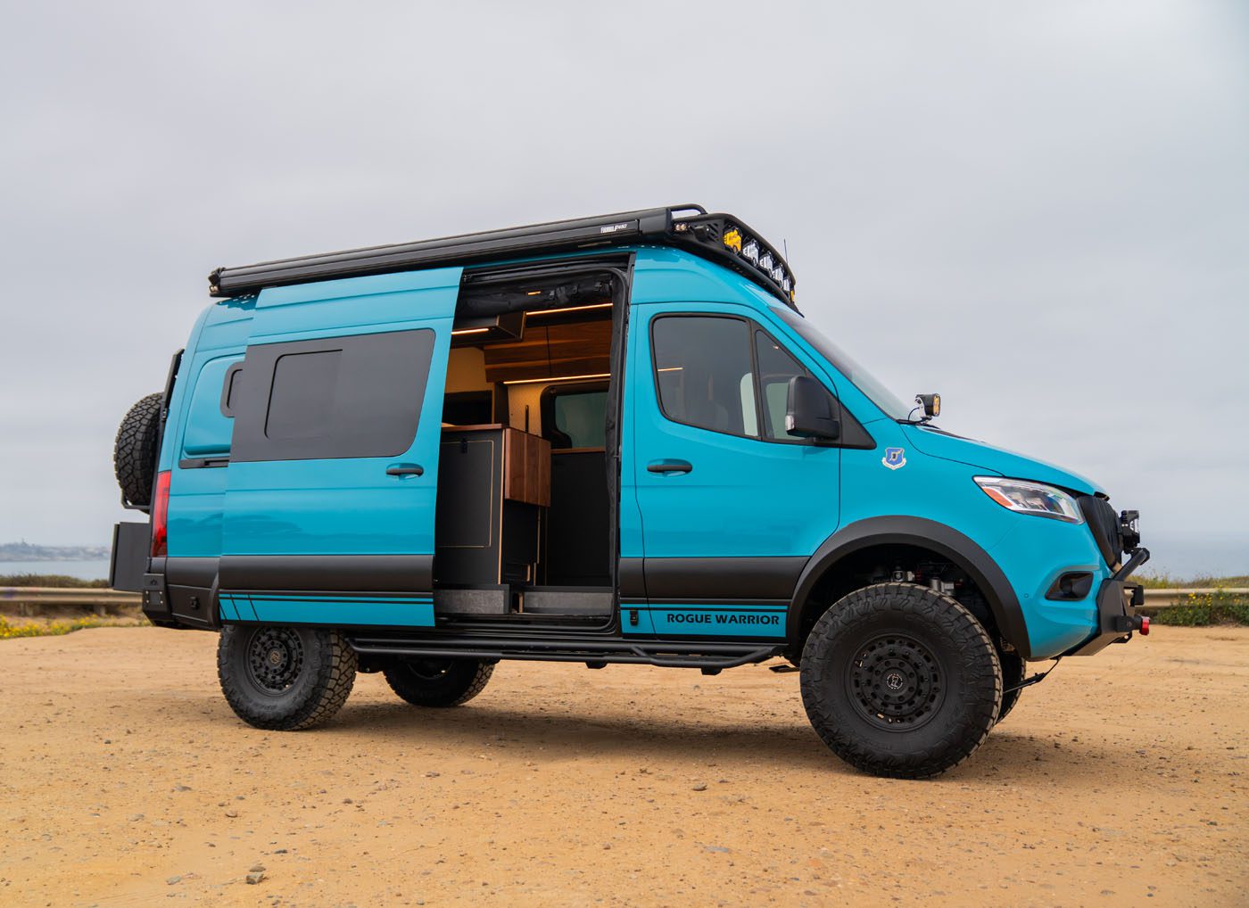 A blue camper van with off-road tires is parked on sandy ground with its side door open, revealing a partial view of the interior.