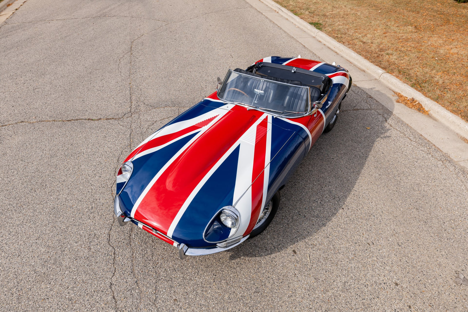 A classic Shaguar, boasting a Union Jack paint design, is parked on a paved road near a grassy area.