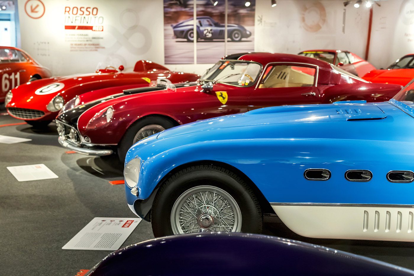A row of classic sports cars, including a blue and several red models, displayed in a well-lit automotive museum exhibit.