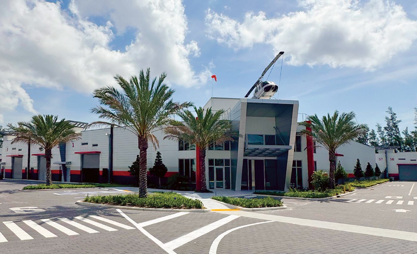 Modern commercial building with palm trees, a glass entrance, and a helicopter mounted on the roof, featuring state-of-the-art storage under a partly cloudy sky.