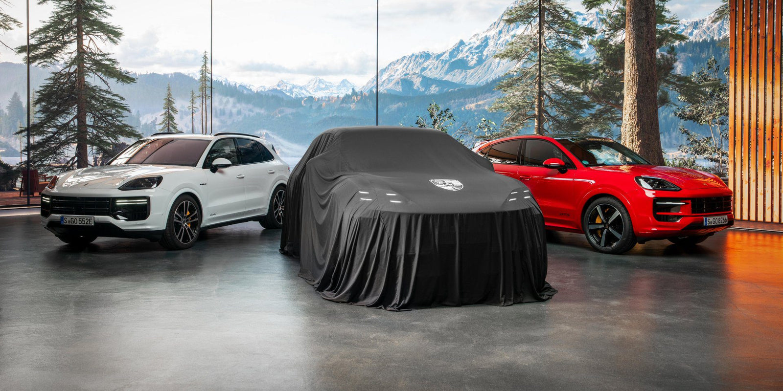 Two Porsche SUVs, a white and a red Cayenne Electric, flank a third vehicle hidden by a black cloth in a Dubai showroom with large windows and mountain views.