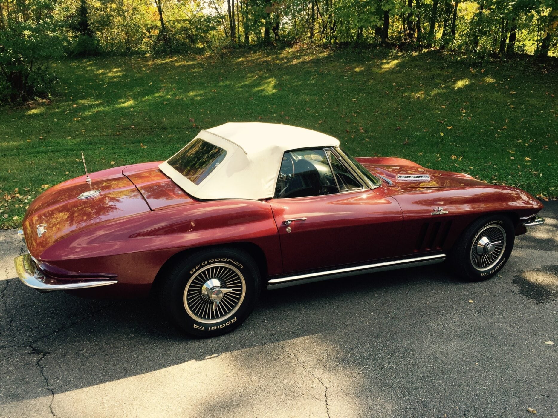 A classic red convertible car with a white soft top is parked on a paved road beside a grassy area with trees, capturing the spirit of the Fall Carlisle Collector Car Auction happening October 2-3, 2025.