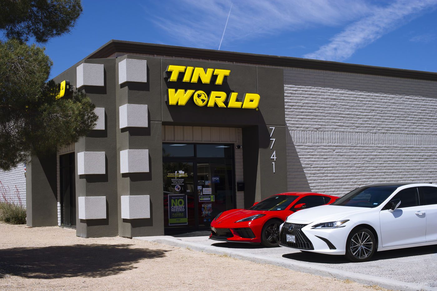 A building with a sign reading "TINT WORLD" above the entrance, with two cars—a red sports car and a white sedan—parked in front.