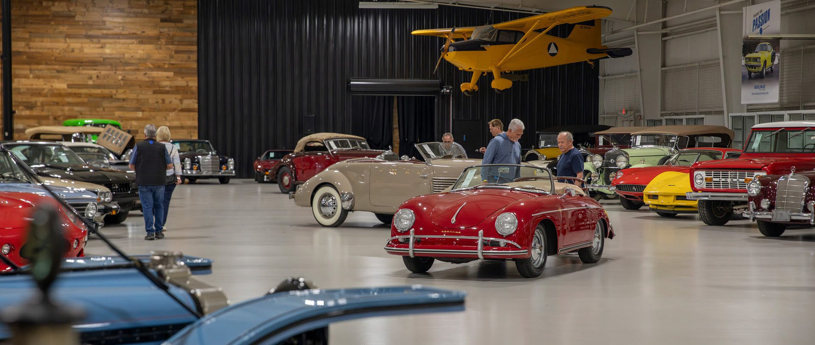 A group of people observe classic cars displayed in a spacious indoor showroom at the Worldwide Auctioneers Enthusiast Auction, with a yellow vintage airplane suspended above.