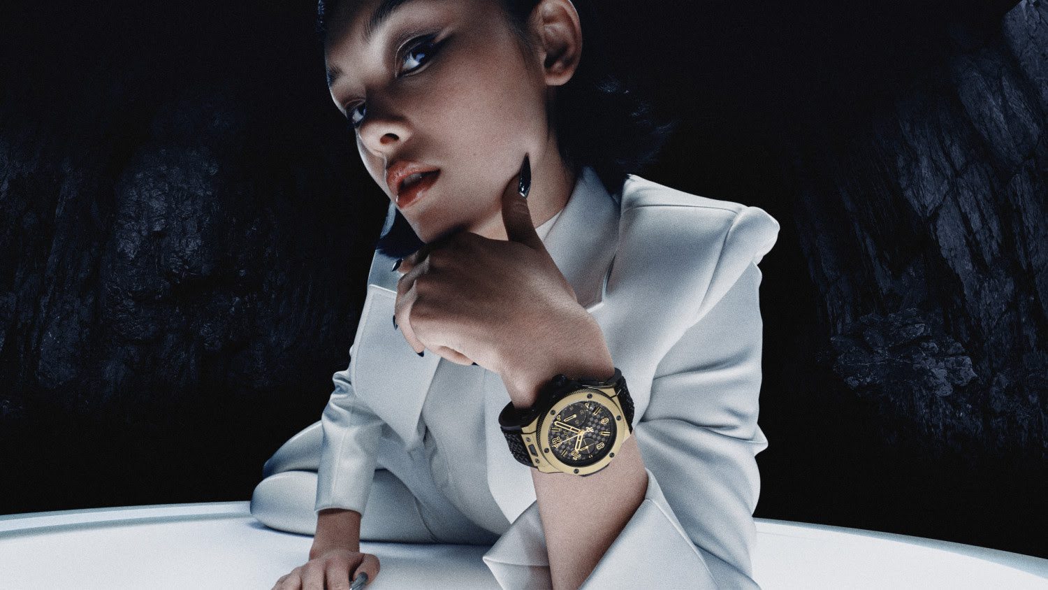 A person in a white suit poses dramatically, resting their chin on one hand, highlighting a large Hublot Big Bang wristwatch. The golden timepiece marks the 20th Anniversary of style against a dark, textured background.