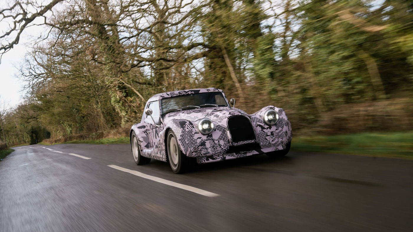 A vintage-style car with camouflage paint drives on a country road lined with leafless trees.
