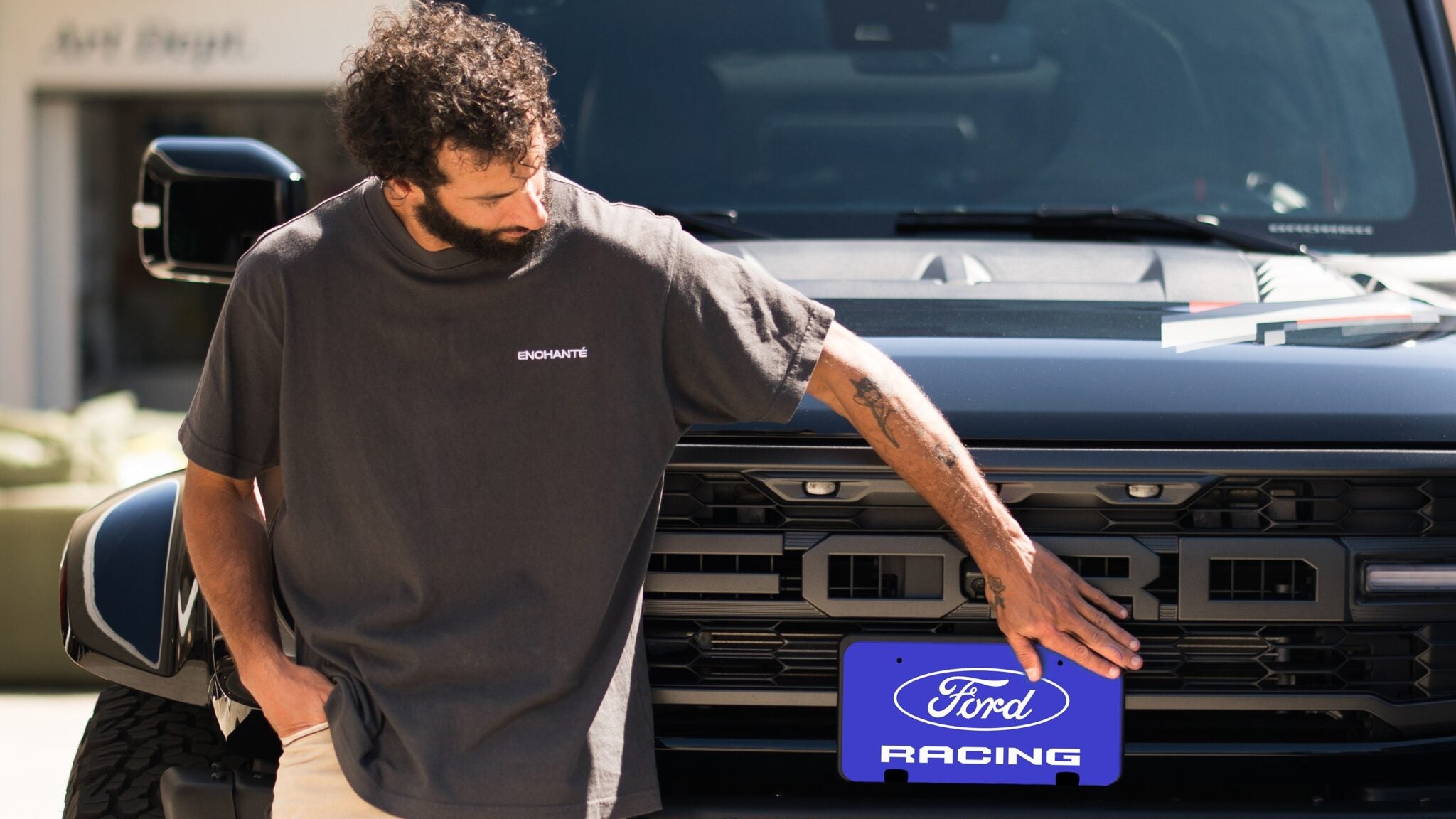 A man places a blue "Ford Racing" license plate on the front of a black Ford truck, proudly showcasing his passion for Ford Performance.