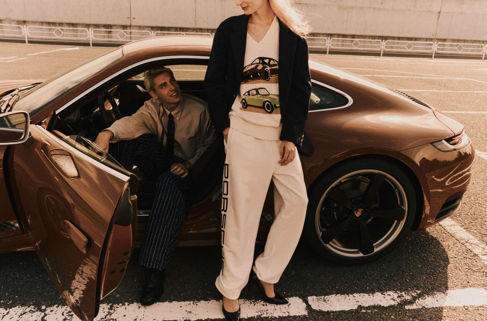 A person sits in the driver's seat of a brown sports car, possibly a Porsche, with the door open, while another stands outside in a Poggy sweater featuring cars, in a parking area.