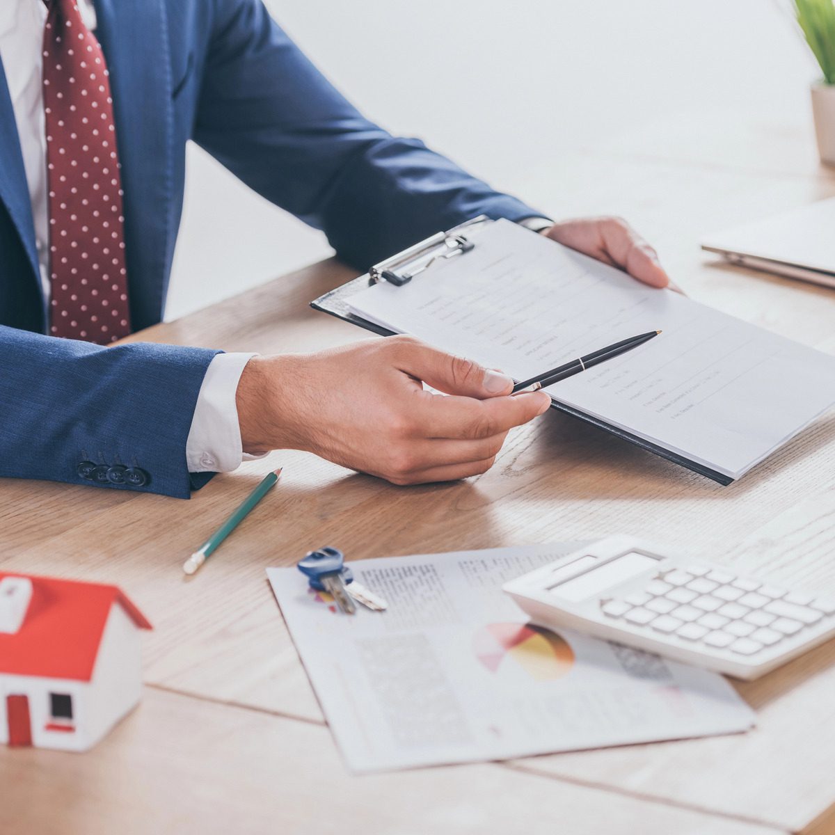 A person in a suit holds a pen and points at a clipboard on a desk with a miniature house, keys, documents, and a calculator, illustrating the personal process of estate settlement.