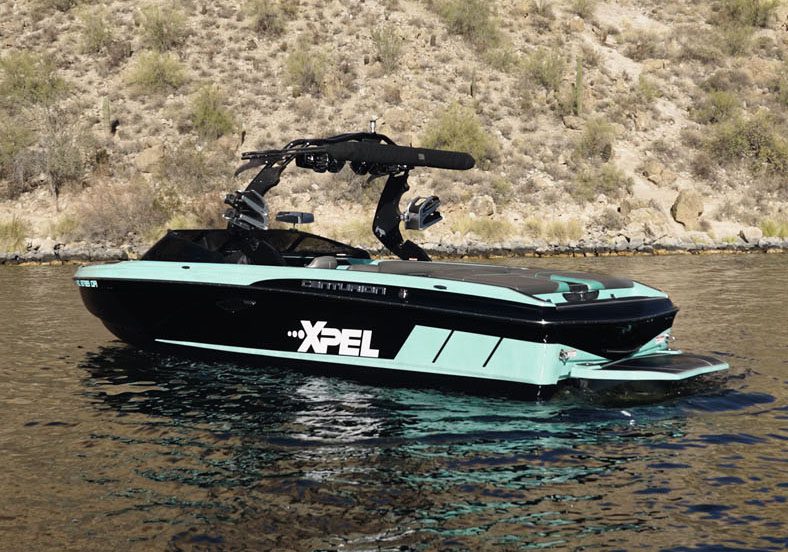 A black and teal motorboat labeled "XPEL" floats on calm water near a rocky, brush-covered shoreline, showcasing the durability of protection films.