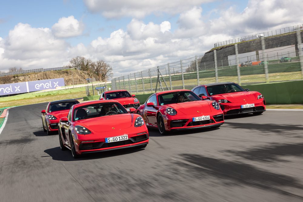 Five red sports cars, symbols of luxury automakers, drive closely together on a racetrack against a backdrop of cloudy skies and fencing.