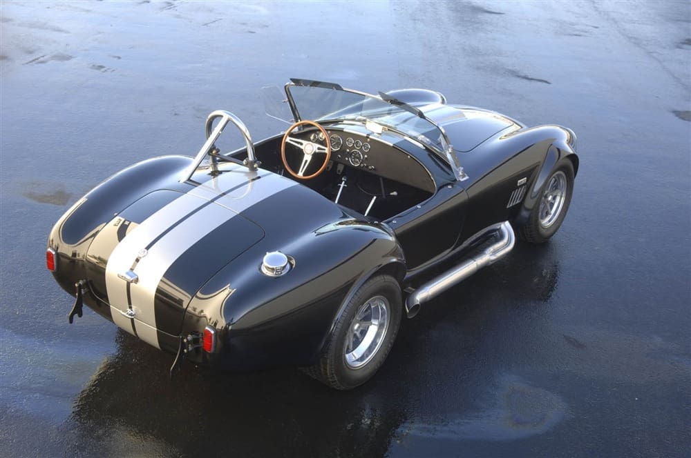 A vintage Shelby 427 Cobra with white racing stripes is parked on wet pavement, featuring an open-top, wood steering wheel, and side exhaust pipes.