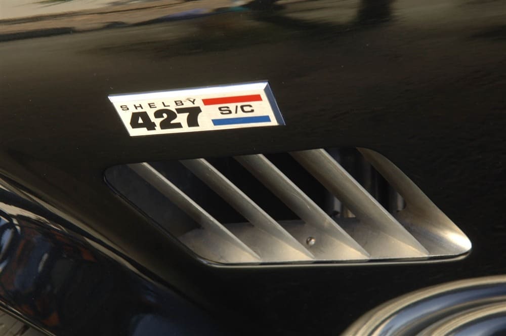 Close-up of a car showcasing a "Shelby 427 Cobra S/C" badge and a vent with horizontal slats.