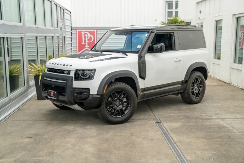 A white Land Rover Defender SUV with black rims is parked on a concrete driveway beside a glass-walled building, showcasing why Land Rover Defenders are considered among the best Land Rover vehicles available.
