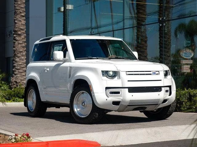 A white Land Rover Defender SUV is parked on a street near a building with palm trees and glass windows, showcasing why many choose to buy Land Rover for both style and adventure.