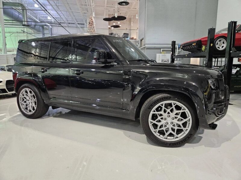 A black Land Rover Defender with tinted windows and silver alloy wheels is parked indoors in a well-lit garage, with other cars visible in the background—an excellent choice for those looking to buy Land Rover vehicles.