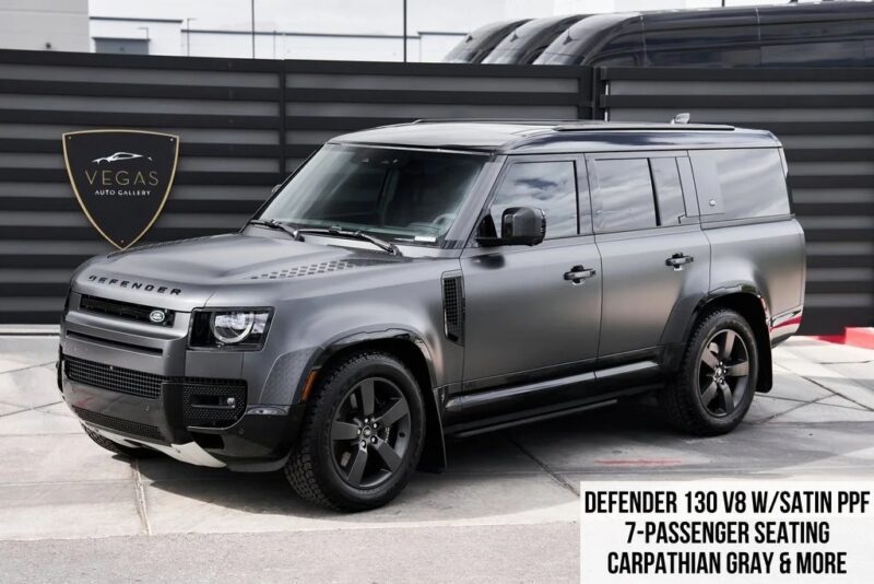 A matte gray Land Rover Defender 130 V8 SUV is parked outside, featuring black wheels and tinted windows. Text overlay highlights its 7-passenger seating and Carpathian Gray color—one of the best Land Rover Defenders you can own.