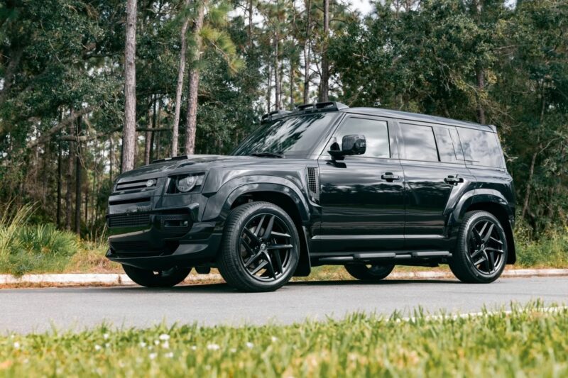 A black SUV, reminiscent of the best Land Rover models, with tinted windows and black rims, is parked on a road surrounded by trees and grass.