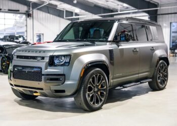 A silver Land Rover Defender SUV is parked indoors in a well-lit showroom, showcasing why it's considered among the best Land Rover vehicles, with other cars visible in the background.