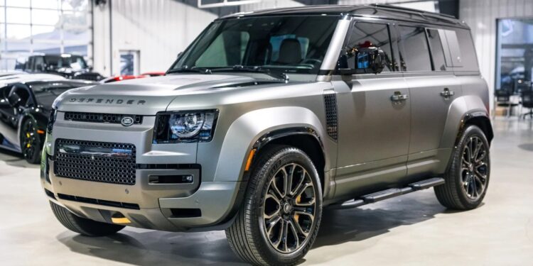 A silver Land Rover Defender SUV is parked indoors in a well-lit showroom, showcasing why it's considered among the best Land Rover vehicles, with other cars visible in the background.