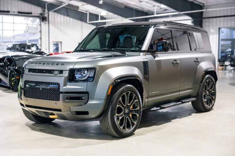 A silver Land Rover Defender SUV is parked indoors in a well-lit showroom, showcasing why it's considered among the best Land Rover vehicles, with other cars visible in the background.