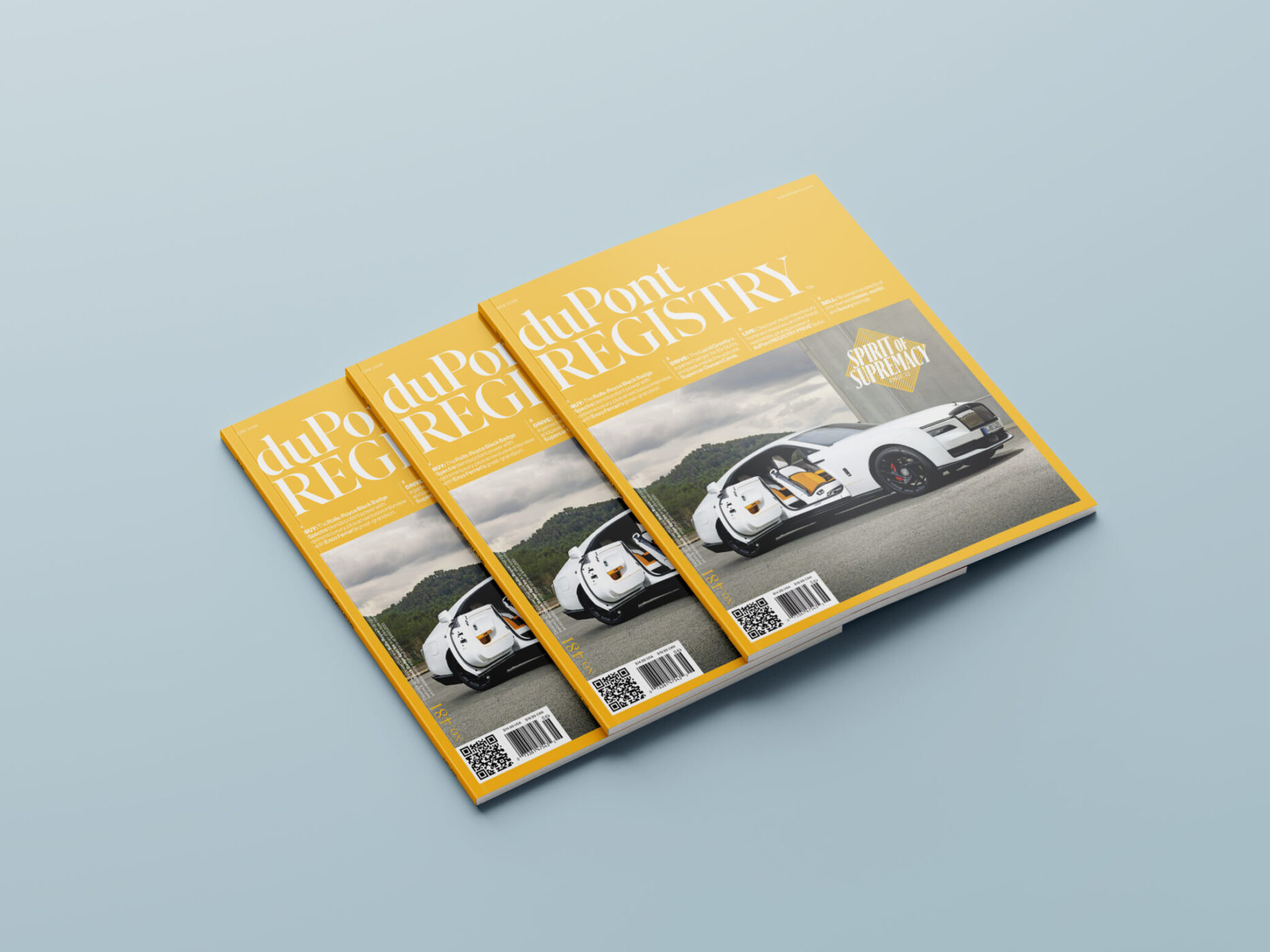 Three copies of the duPont Registry magazine with a yellow cover featuring a white sports car on a winding road, placed on a light blue surface.
