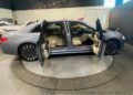 The rare Lincoln Continental, Coach Door Edition, is displayed with all four doors open, revealing a chic beige interior. It sits elegantly on a circular platform inside the showroom, highlighting its silver luxury.