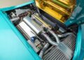 Under the teal exterior of this 1993 Vector Avtech WX-3, a 1-of-1 masterpiece, lies a car engine compartment with exposed metal components, including pipes, a radiator, and a silver engine cover. Some areas feature gold insulation. Now available for sale for discerning collectors.