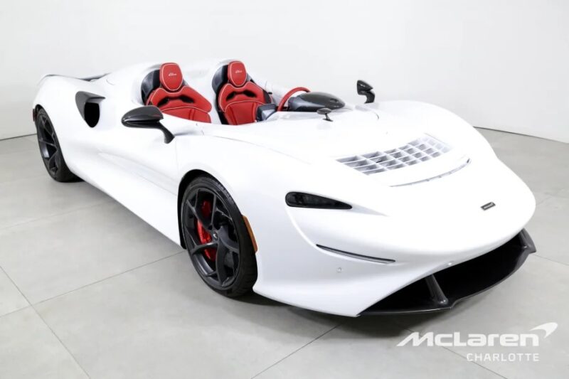 A sleek white McLaren Elva, the ultimate ultra-light thrill machine with red accents, no windshield, and a modern aerodynamic design, is parked indoors on a gray floor.