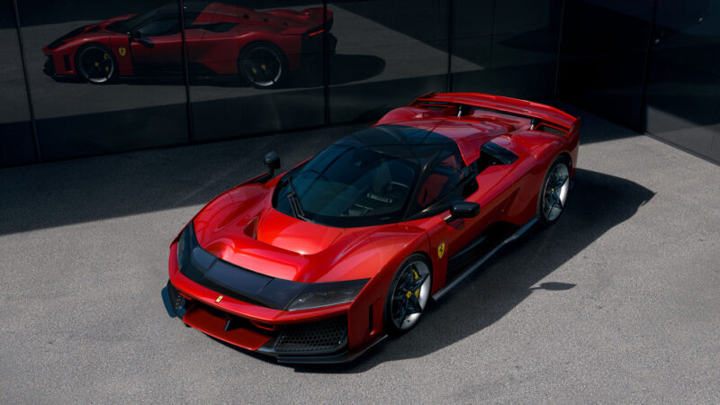 A sleek red sports car, part of the exciting 2024 cars lineup, is parked on a gray surface near a reflective glass wall, where its reflection adds to the allure.