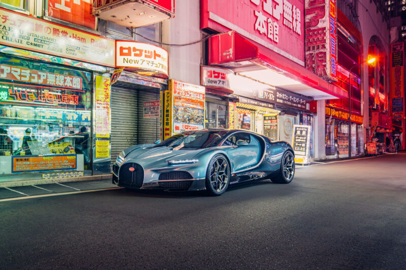 A sleek sports car, part of the dazzling 2024 lineup, is parked on a brightly lit street, surrounded by vibrant storefronts with neon signs and advertisements in a city setting.