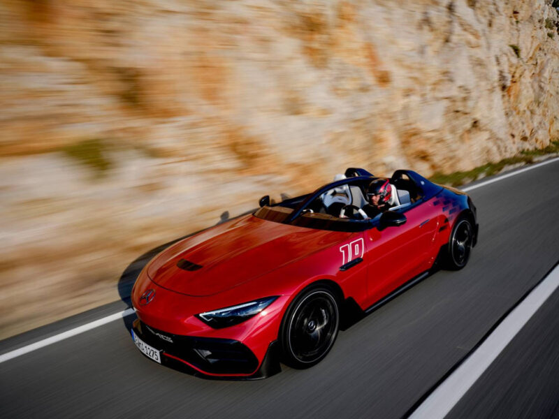 The 2024 red convertible sports car, newly revealed with sleek black accents, speeds along a coastal road, mountains majestically framing the background.