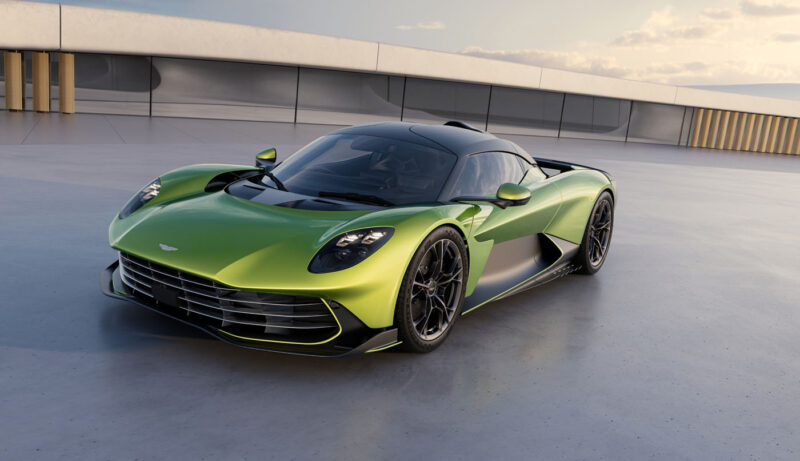 A sleek green sports car with a black roof is parked on a smooth concrete surface, poised under a cloudy sky, with the modern building in the background hinting at an exclusive 2024 car reveal.