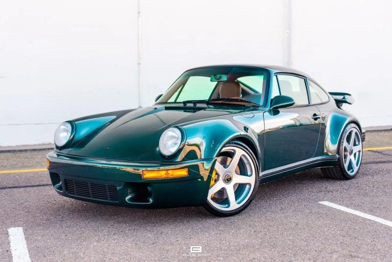 A classic dark green sports car with a sleek design and five-spoke alloy wheels, reminiscent of St. Patrick's Day charm, parked on a paved surface in front of a white wall.