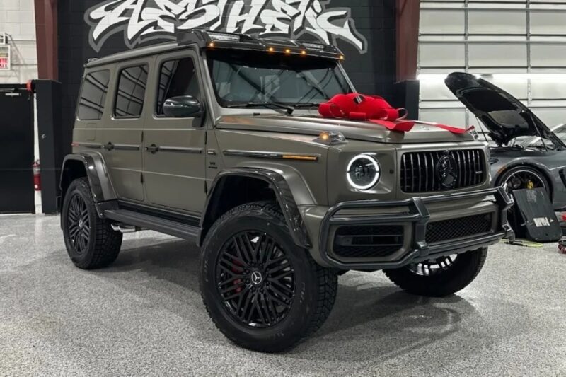 A gray luxury SUV with off-road tires is parked indoors, adorned with a red bow on the hood. Behind it, a stylized logo is set against a wall adorned with subtle hints of green, adding a touch of St. Patrick's Day charm to the scene.