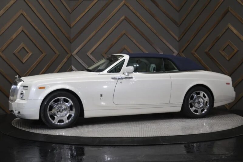 A white convertible luxury car with a blue soft top, reminiscent of a Rolls-Royce Phantom, is displayed on a rotating platform against a geometric-patterned wall.