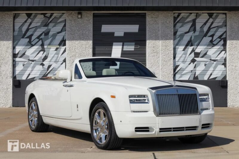 A white convertible Rolls-Royce Phantom, the epitome of quintessential luxury, is elegantly parked in front of a building with patterned windows.