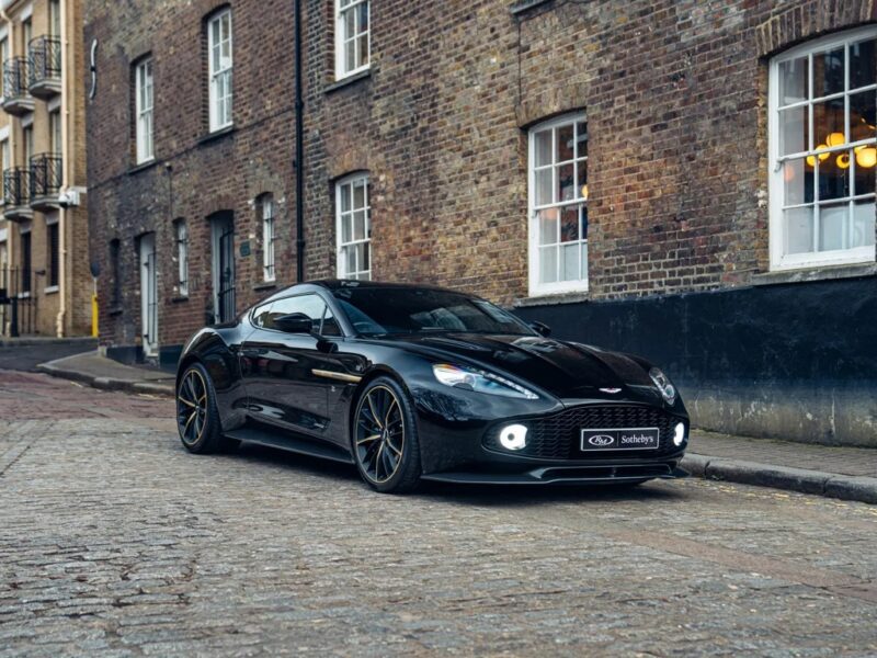 A black Aston Martin Vanquish is parked on a cobblestone street beside a brick building, turning heads with its striking design.
