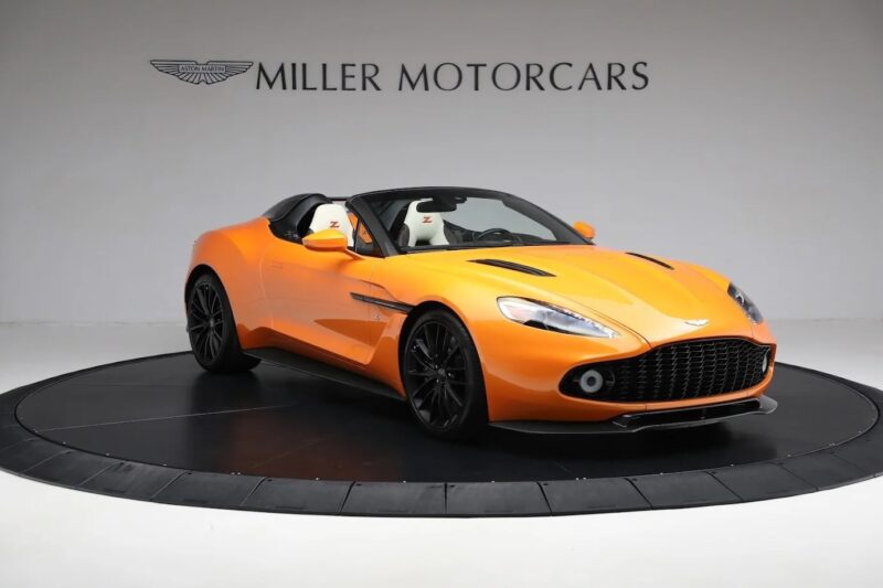An orange Aston Martin Vanquish convertible sports car is displayed on a black turntable in a showroom with "Miller Motorcars" on the wall in the background, highlighting this stunning model as a premier luxury vehicle for sale.