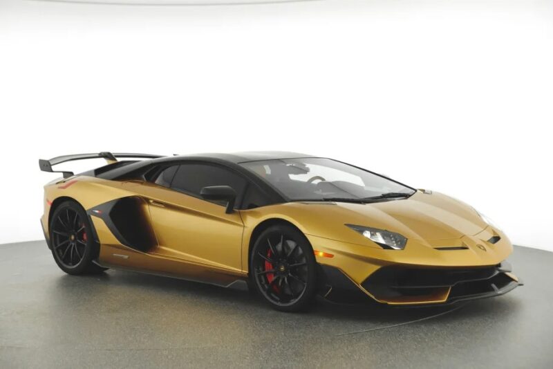 A gold Lamborghini Aventador SVJ with black accents is parked in a well-lit studio. It features a sleek design, large rear wing, and black wheels with red brake calipers. Ideal for those looking to buy the best Lamborghini.