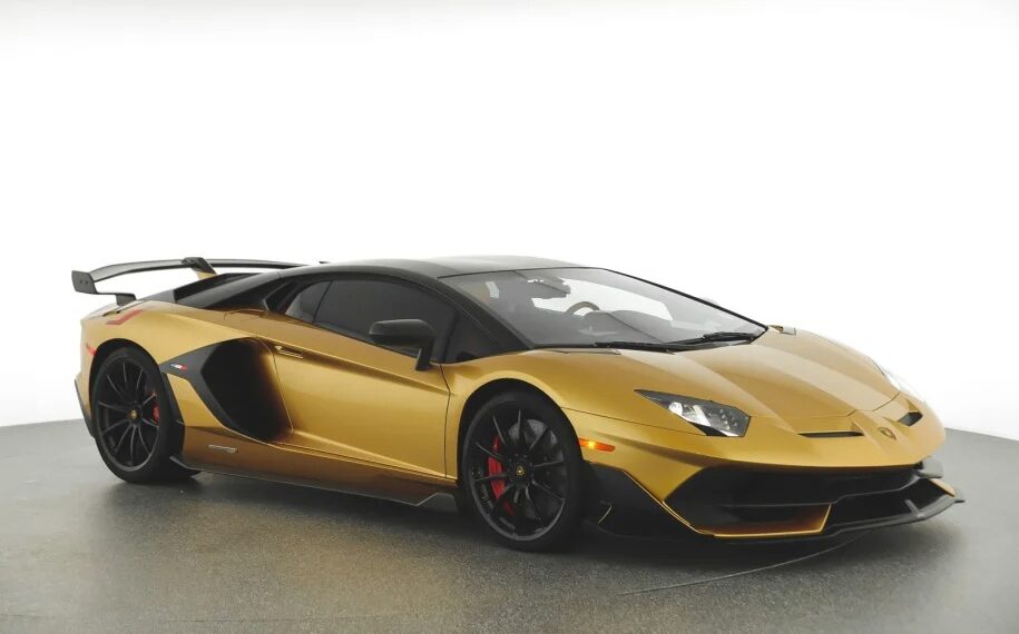 A gold Lamborghini Aventador SVJ with black accents is parked in a well-lit studio. It features a sleek design, large rear wing, and black wheels with red brake calipers. Ideal for those looking to buy the best Lamborghini.