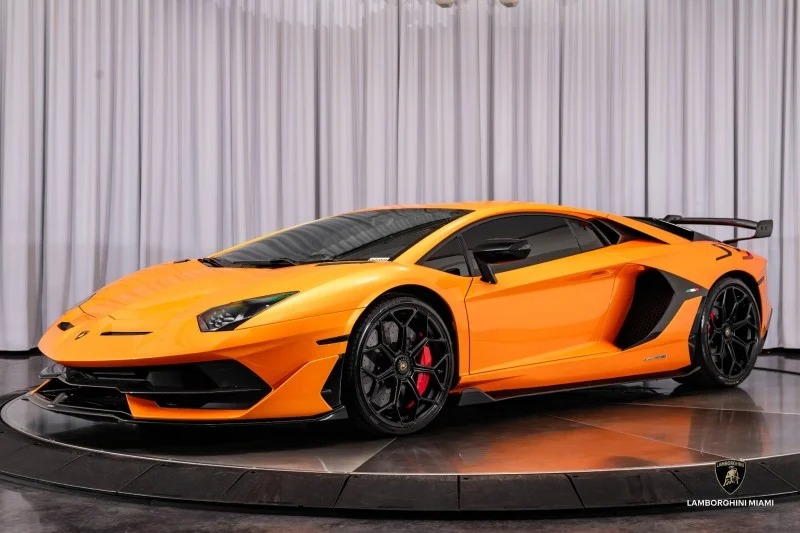 The sleek Lamborghini Aventador SVJ, in a vibrant orange hue, is showcased indoors on a turntable against a white curtain backdrop.