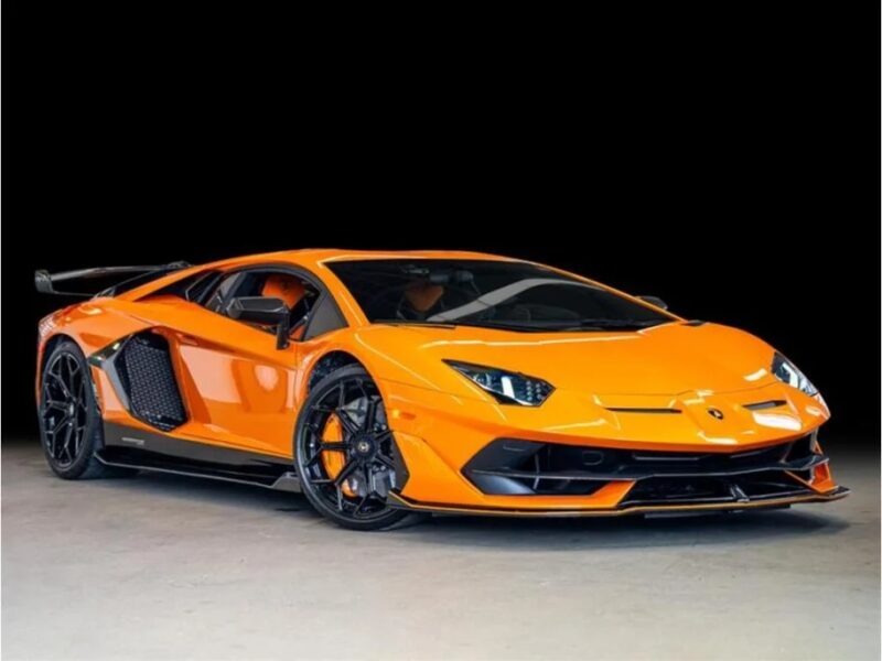 Today, an orange Lamborghini Aventador SVJ with aerodynamic design and black accents sits sleekly atop a gray surface against a black background, enticing car enthusiasts to consider their next buy.