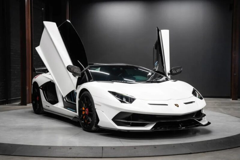 A white Lamborghini Aventador with butterfly doors open is showcased indoors on a circular platform, illuminated softly in the dimly lit room for an exclusive display.