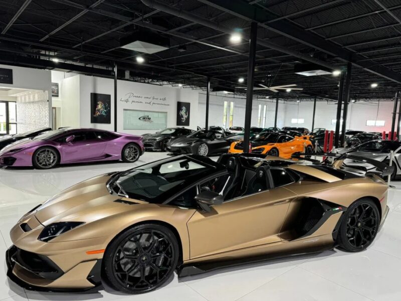 A showroom filled with luxury sports cars, highlighted by a gold convertible Lamborghini Aventador SVJ in the foreground, surrounded by various other stunning models in the background.