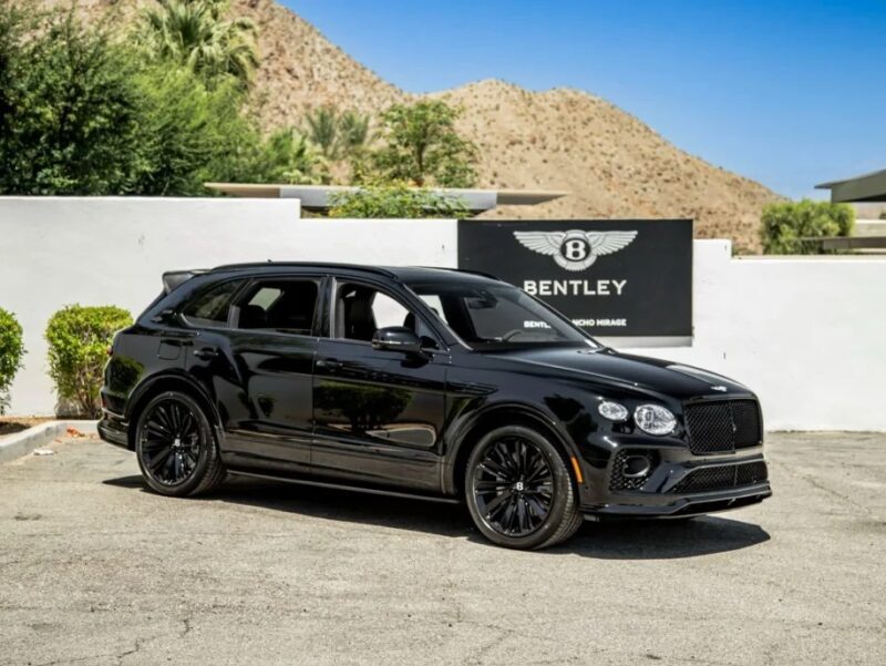 A luxury black Bentley SUV is parked in front of a Bentley dealership sign, set against a breathtaking desert landscape and clear blue sky, embodying performance at its finest.